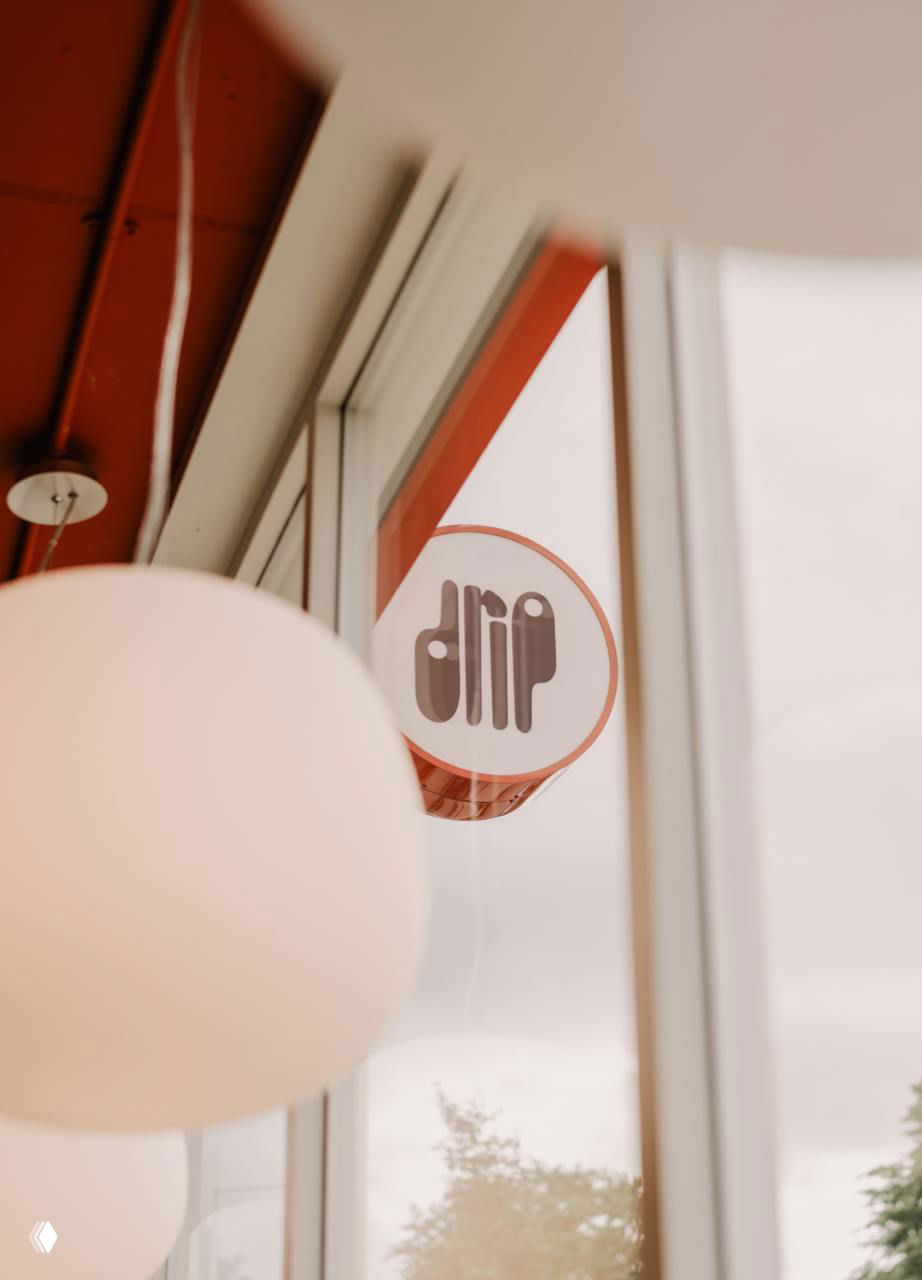 Круглый знак Drip виден в отражении витрины изнутри кафе, передний план занимает мягкий белый светильник; кадр подчёркивает атмосферу и визуальную идентичность.