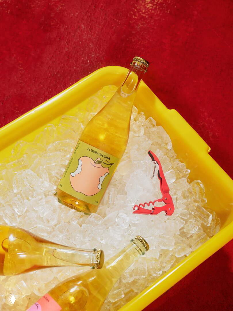 Бутылка сидра в ящике со льдом на красном фоне рядом с открывалкой, цветной и экспрессивный художественный кадр оформления продукта.