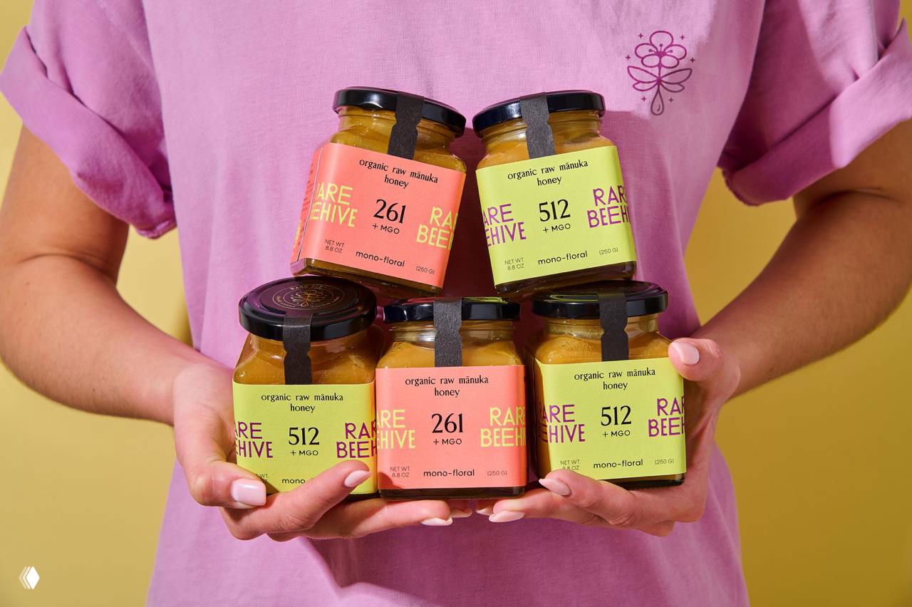 Человек в розовой футболке держит в руках несколько баночек Rare Beehive, демонстрируя размеры и сочетание коралловых и лимонных этикеток в стопке.