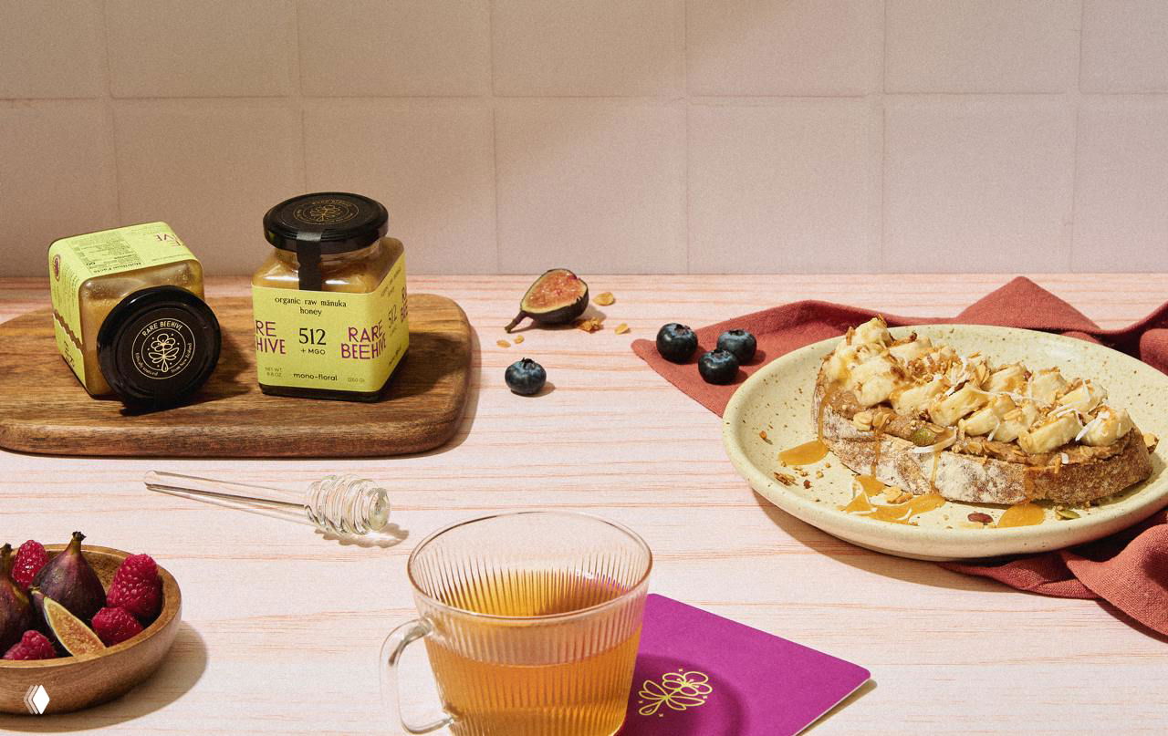 Стилизованная натюрморт-сцена с баночками Rare Beehive, чашкой чая, тостом с мёдом и ягодами — показ потребительского контекста продукта.