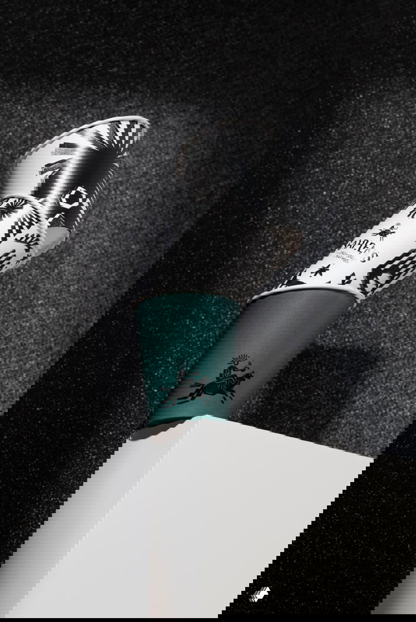 Стопка бумажных стаканов Sapiens с чёрно‑белыми линогравюрами и зелёным однотонным стаканом на мелкозернистом тёмном фоне.