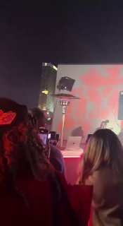 Зрители на представлении снимают сцену мобильными телефонами; на заднем плане видна световая проекция и экран шоу дронов.