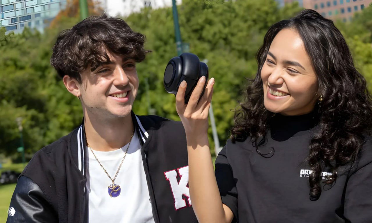 Двое улыбающихся людей на улице рассматривают модуль наушников TDM в руках; демонстрация складной конструкции и функции превращения в колонку.