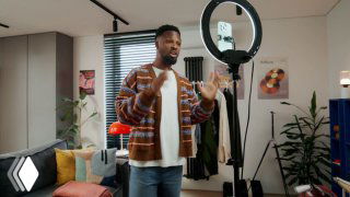 Мужчина в домашней студии перед кольцевой лампой и камерой, снимает видео на фоне мебели и растений, показывает оборудование.