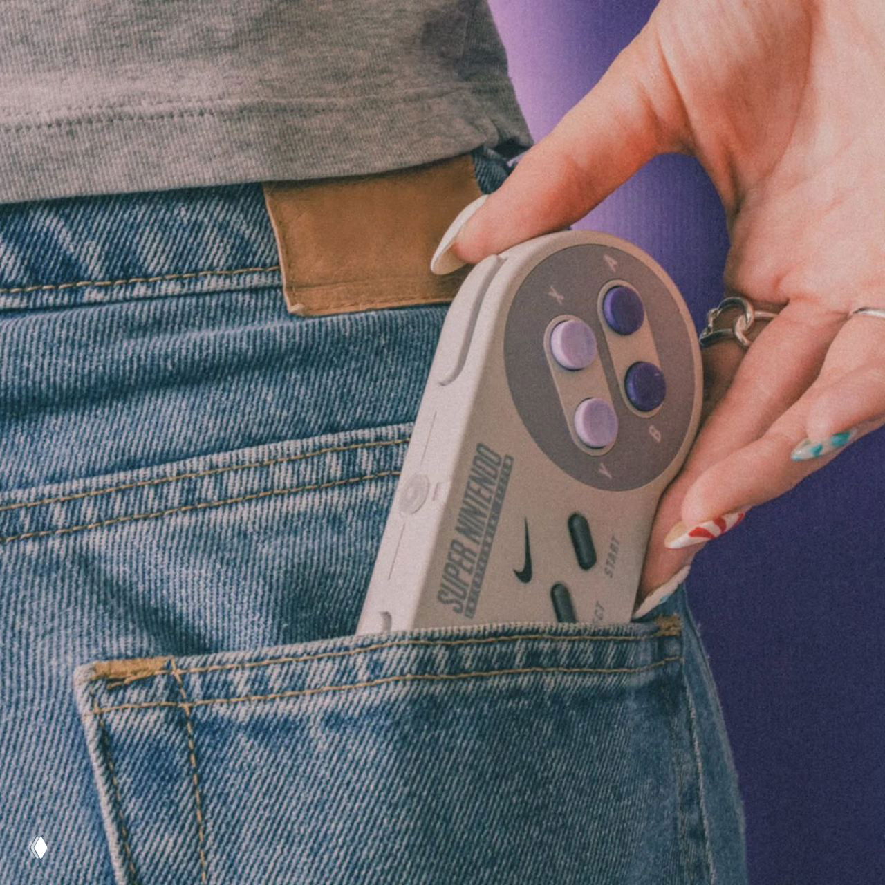 Рука достаёт компактный контроллер Super Nintendo с логотипом Nike из заднего кармана джинсов — крупный план, текстура ткани и маникюр.