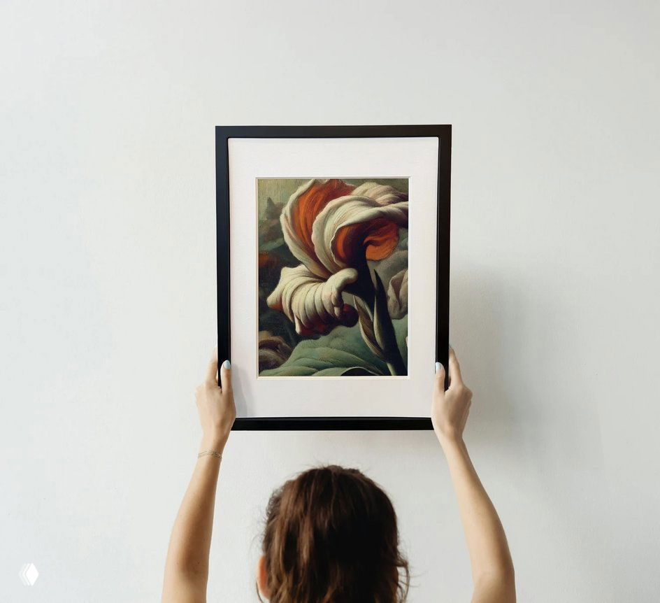 Крупный план: человек поднимает рамку с изображением цветка на фоне белой стены, видны руки и верхняя часть головы, демонстрация произведения в раме.