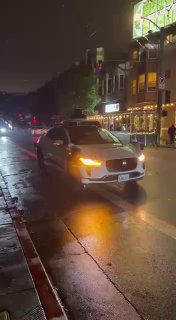 Близкий план автономного такси Waymo на пустой ночной улице Сан‑Франциско; фары и габаритные огни отражаются в лужах при отключении электричества.