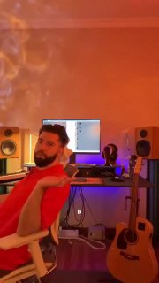 Широкий кадр: мужчина в кресле демонстрирует студийное рабочее место — стол с компьютером, гитара у стены и акустические мониторы, тёплое освещение.