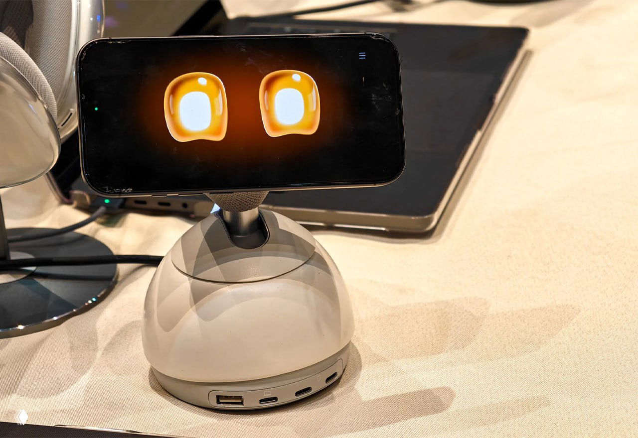 Демонстрация DeskMate на выставочном стенде CES: iPhone в держателе над куполообразной базой с портами, на экране видны мультяшные глаза.