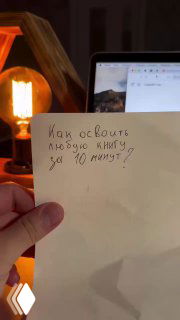 Фото рукописной заметки на листе с фразой «Как освоить любую книгу за 10 минут?», рука держит лист, на заднем плане ноутбук и настольная лампа