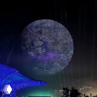 Ночное световое шоу: тысячи дронов в небе над городом формируют большую круглую светящуюся проекцию на фоне городской подсветки и горизонта.