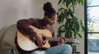 Человек в комнате играет на акустической гитаре и носит современные очки; кадр показывает повседневное использование носимого устройства в интерьере, естественное освещение.