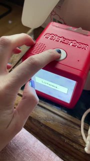 Красный компактный принтер Stickerbox на деревянной поверхности: крупный план кнопки и маленького экрана, на экране видно слово «Listening…», рука готовится нажать кнопку.