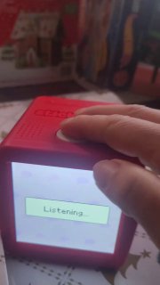 Ещё крупный план красного корпуса Stickerbox с подсветкой экрана и надписью «Listening…», видна рука пользователя и праздничный фон на столе.