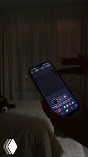 Рука держит смартфон с открытым приложением управления подсветкой на фоне затемнённой комнаты, занавесок и кровати — демонстрация управления гирляндой через телефон.