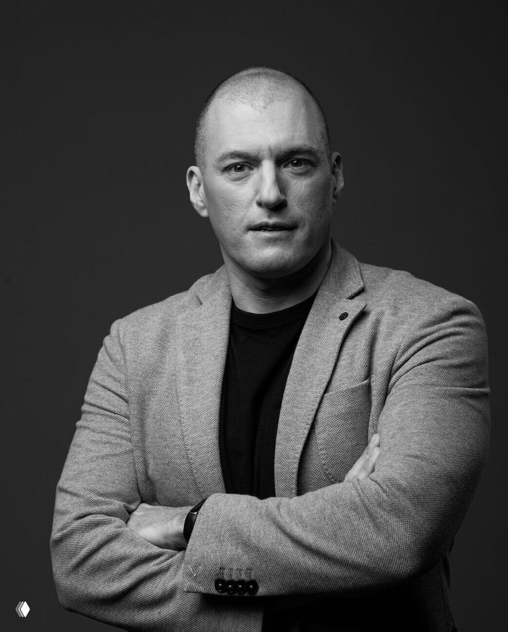 Черно‑белый студийный портрет мужчины в пиджаке с перекрещенными руками, деловой образ на нейтральном фоне, серьезный ракурс.