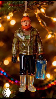 Крупный план стеклянной елочной игрушки в виде фигурки человека в зимней куртке, подвешенной к еловой ветке на фоне тёплого праздничного боке.