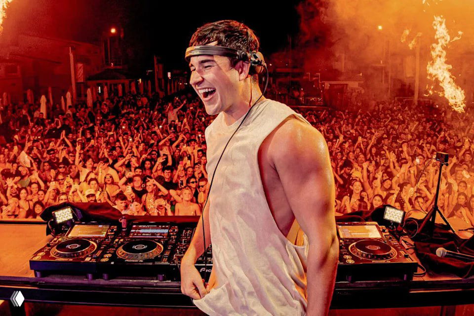 Диджей на сцене перед большим восторженным толпой на ночном концерте, артист улыбается за пультом, яркие огни и эффектные декорации вокруг