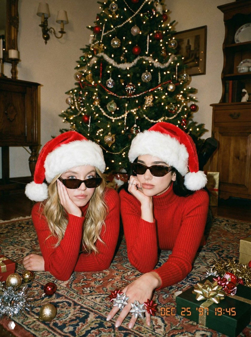 Две молодые женщины в красных свитерах и шапках Санты сидят на ковре у наряженной ёлки, расслабленная поза и мягкое боке на заднем плане, винтажная эстетика.
