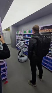 Робот-мерчандайзер в торговом проходе сортирует и раскладывает товары по полкам; рядом человек снимает сцену, видны стеллажи и упаковки.