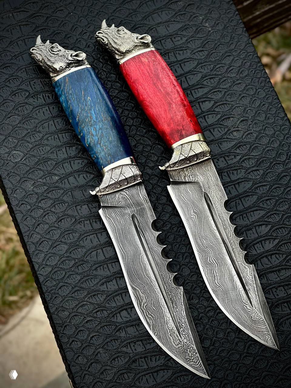 Пара декоративных ножей крупным планом: рисунок дамаска на лезвии, мельхиорная гарда и красная/синяя рукояти из карельской берёзы.