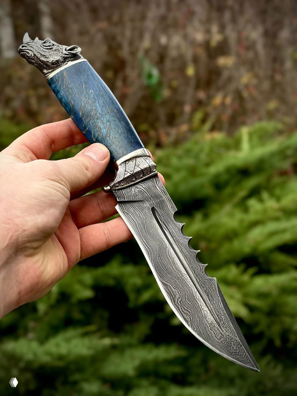 Нож в руке крупным планом: дамасковый клинок с декоративной выточкой и синяя рукоять из карельской берёзы, мельхиорная гарда видно ясно.