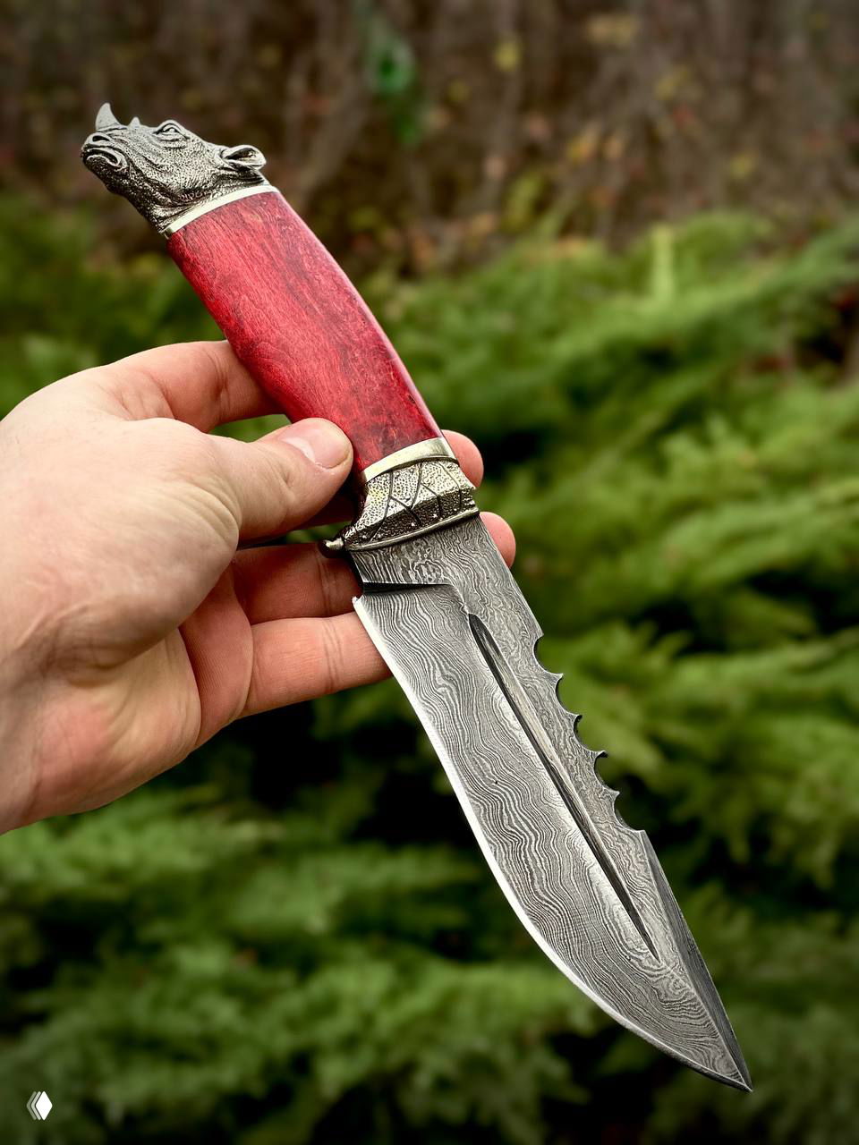 Нож в руке с красной рукоятью из карельской берёзы и дамасковым лезвием, фон — зелёная растительность на улице.