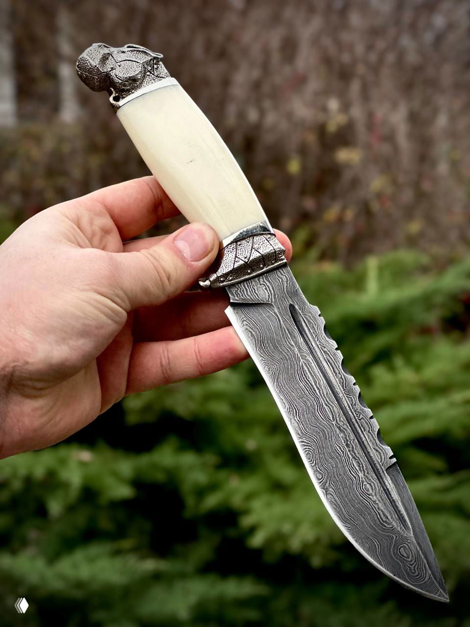 Крупный план ножа в руке: узор дамасской стали на клинке, светлая акриловая рукоять и мельхиоровая гарда на фоне природы