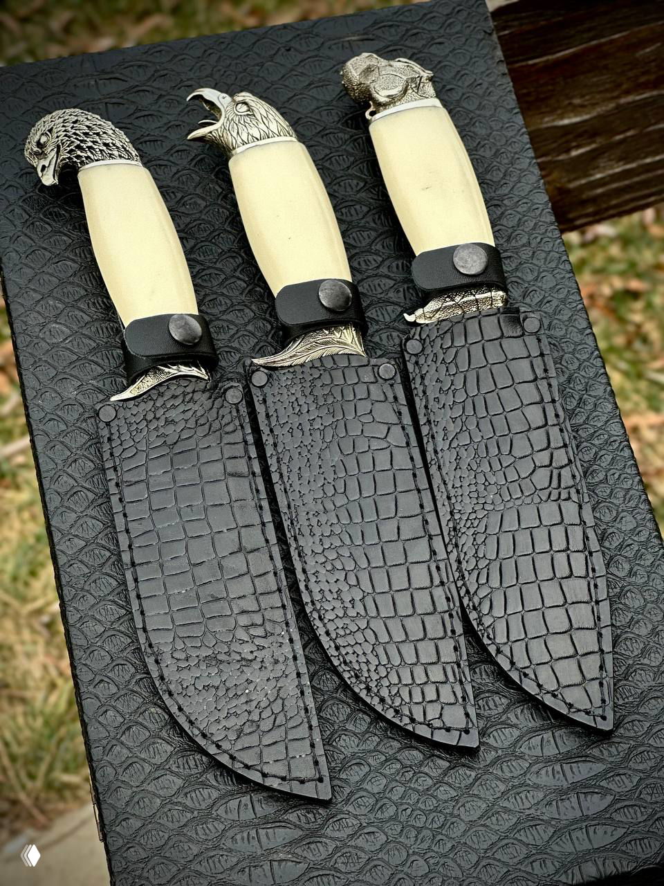 Три ножа в кожаных чёрных ножнах с тиснением, расположенные на текстурированной подложке, видны детали швов и застёжек