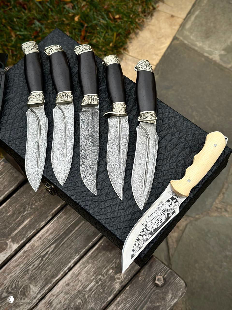 Крупный план пяти дамасских клинков с декоративными гардой и одного ножа со светлой резной рукоятью на текстурированном футляре