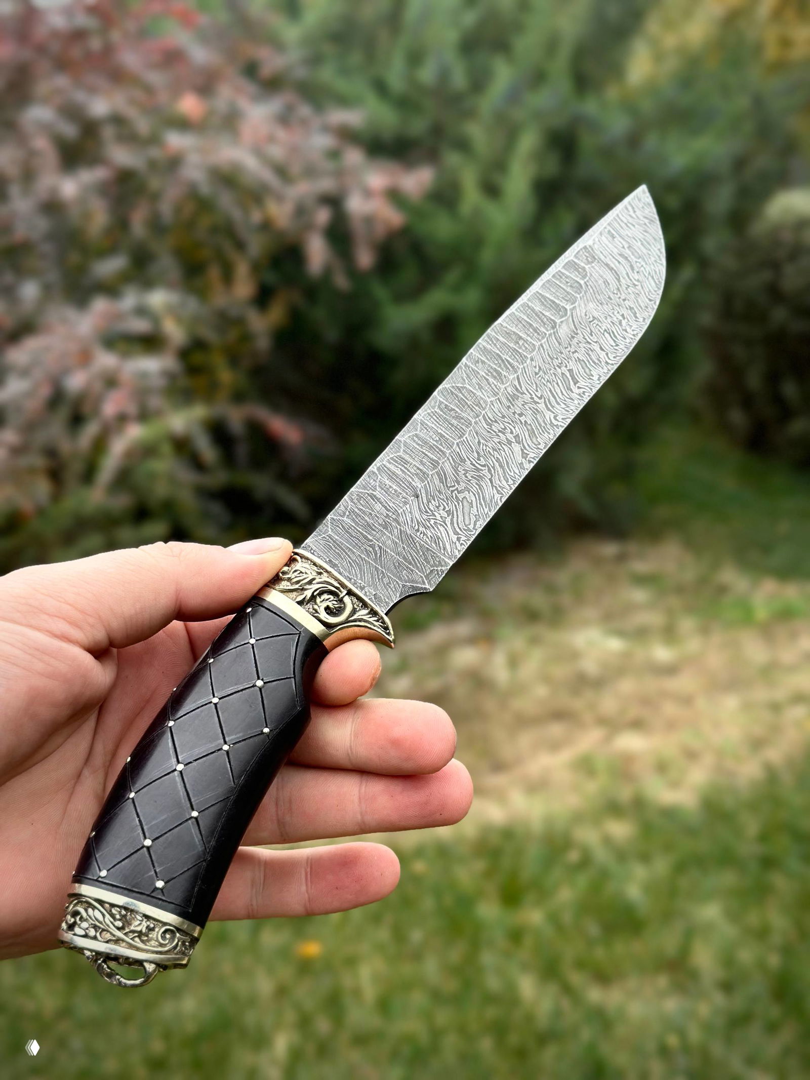 Фотография длинного дамасского клинка под углом на размытом зелёном фоне, хорошо видна текстура и профиль лезвия.