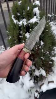 Крупный кадр ножа в руке с боковым ракурсом клинка: выраженный дамасский рисунок, видна форма обуха и острие на фоне хвойного куста со снегом.