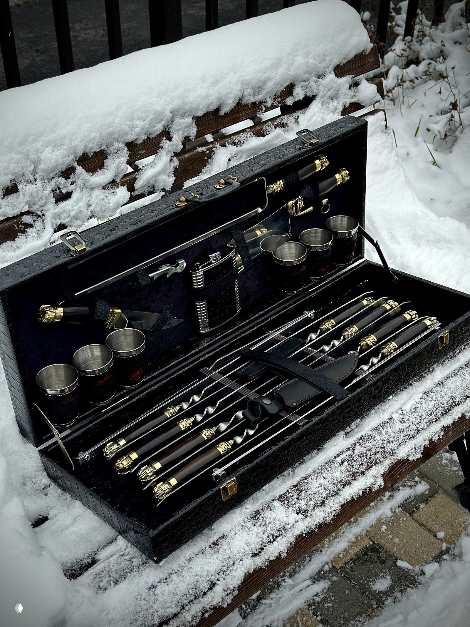 Крупный план внутренней укладки набора: ножи, топор, фляжка и металлические стопки зафиксированы тканевыми ремнями в бархатной выстилке кейса.