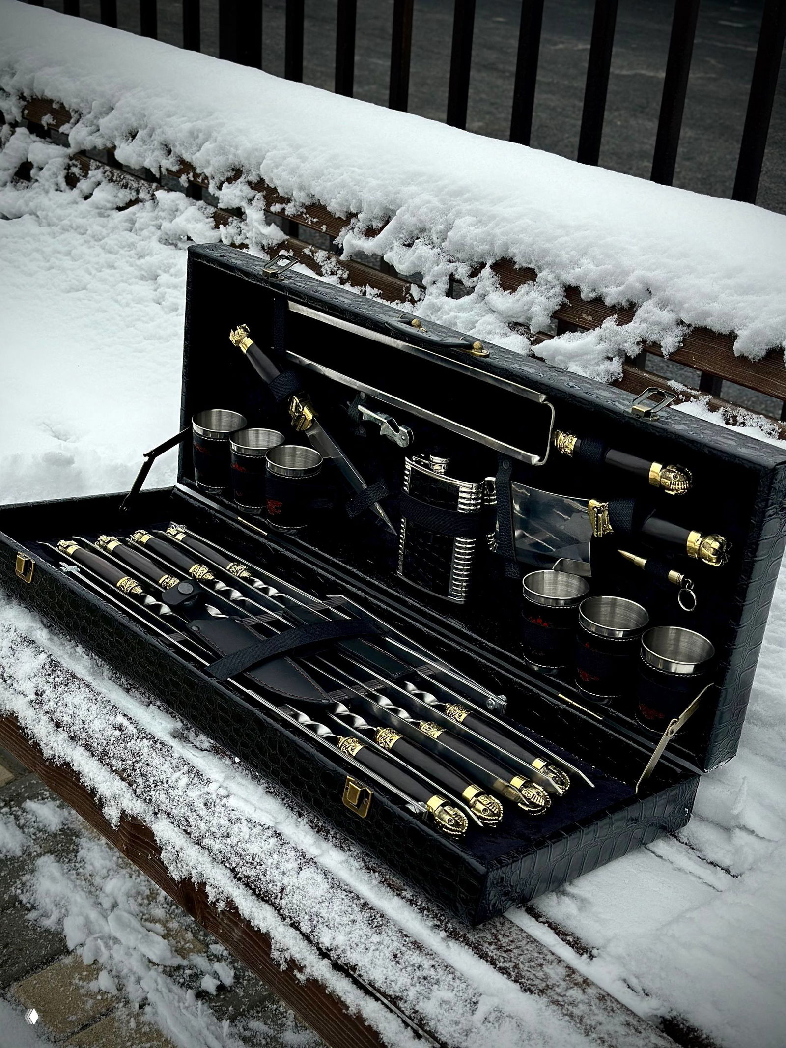 Чемодан‑кейc с набором для шашлыка на снежной скамье: видны рисунок кожаной обложки, металлические пряжки и выдвинутые элементы комплекта.
