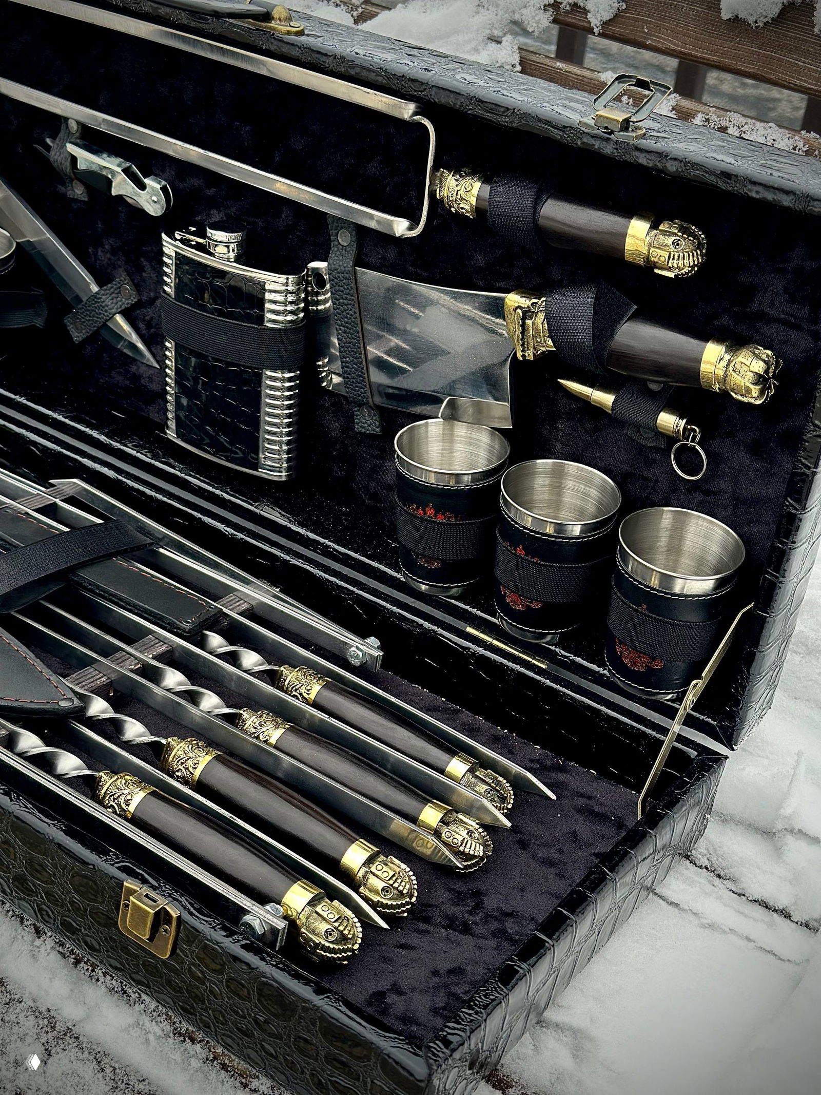 Вид на ряд шампуров с декоративными латунными наконечниками и кожаными ручками, уложенных в нижней части кейса на фоне снега.