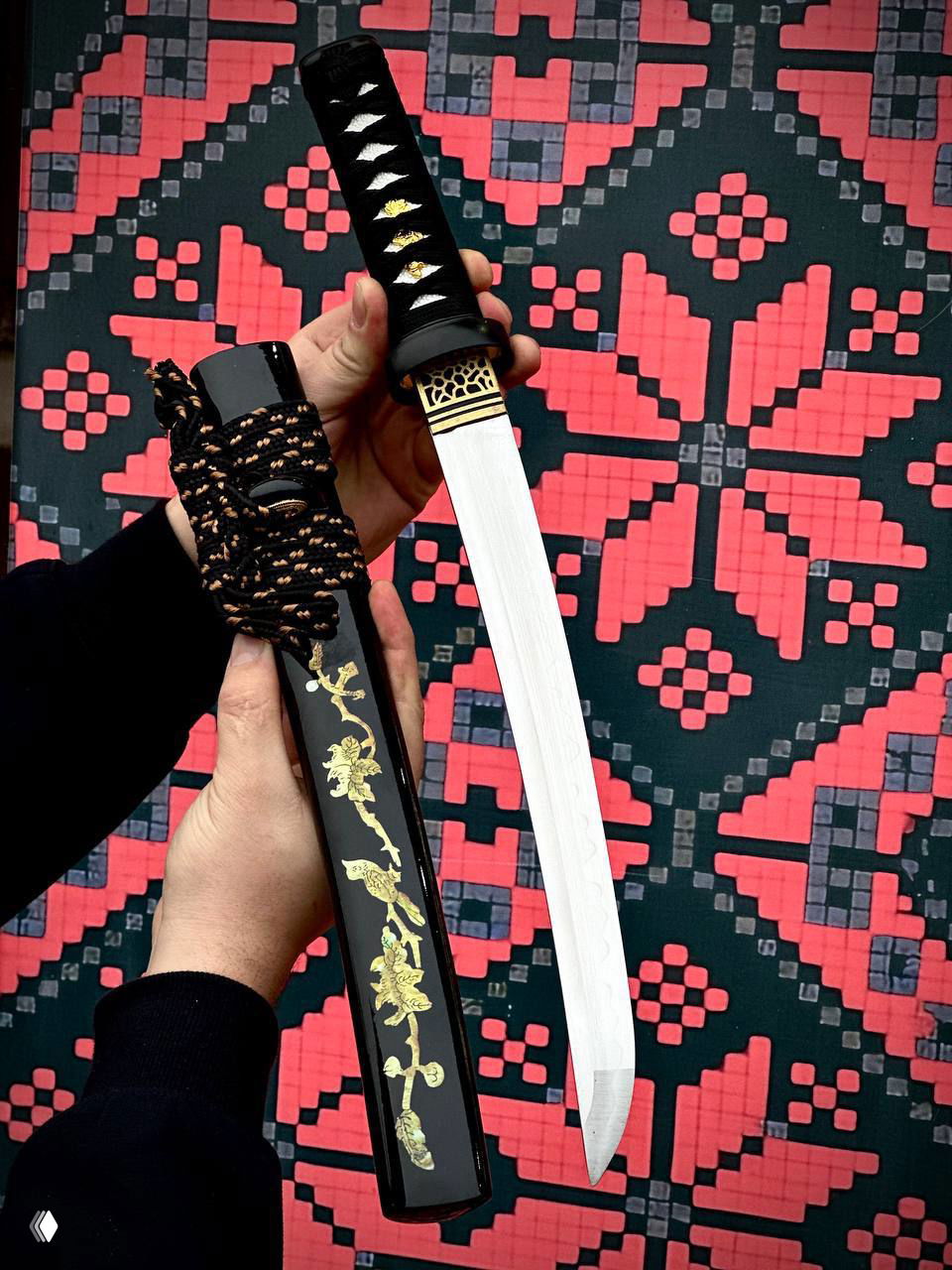 Вакидзаси со светлым клинком и лакированными ножнами в руке мастера, видно золотой орнамент и переплёт тесаки на рукояти.