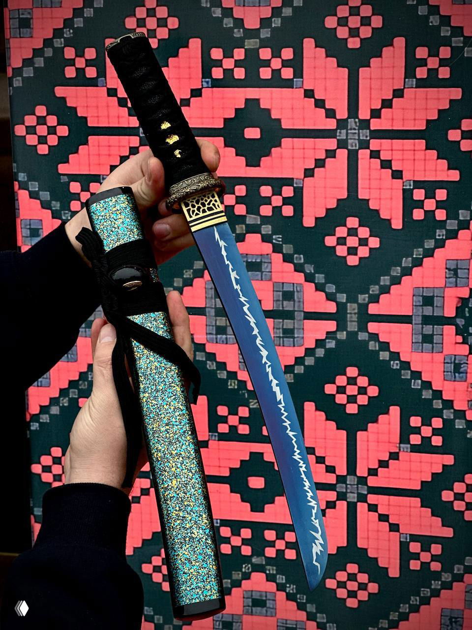 Синяя вариация вакидзаси с декоративным рисунком на клинке и блестящими бирюзовыми ножнами в руках мастера на узорной подложке.