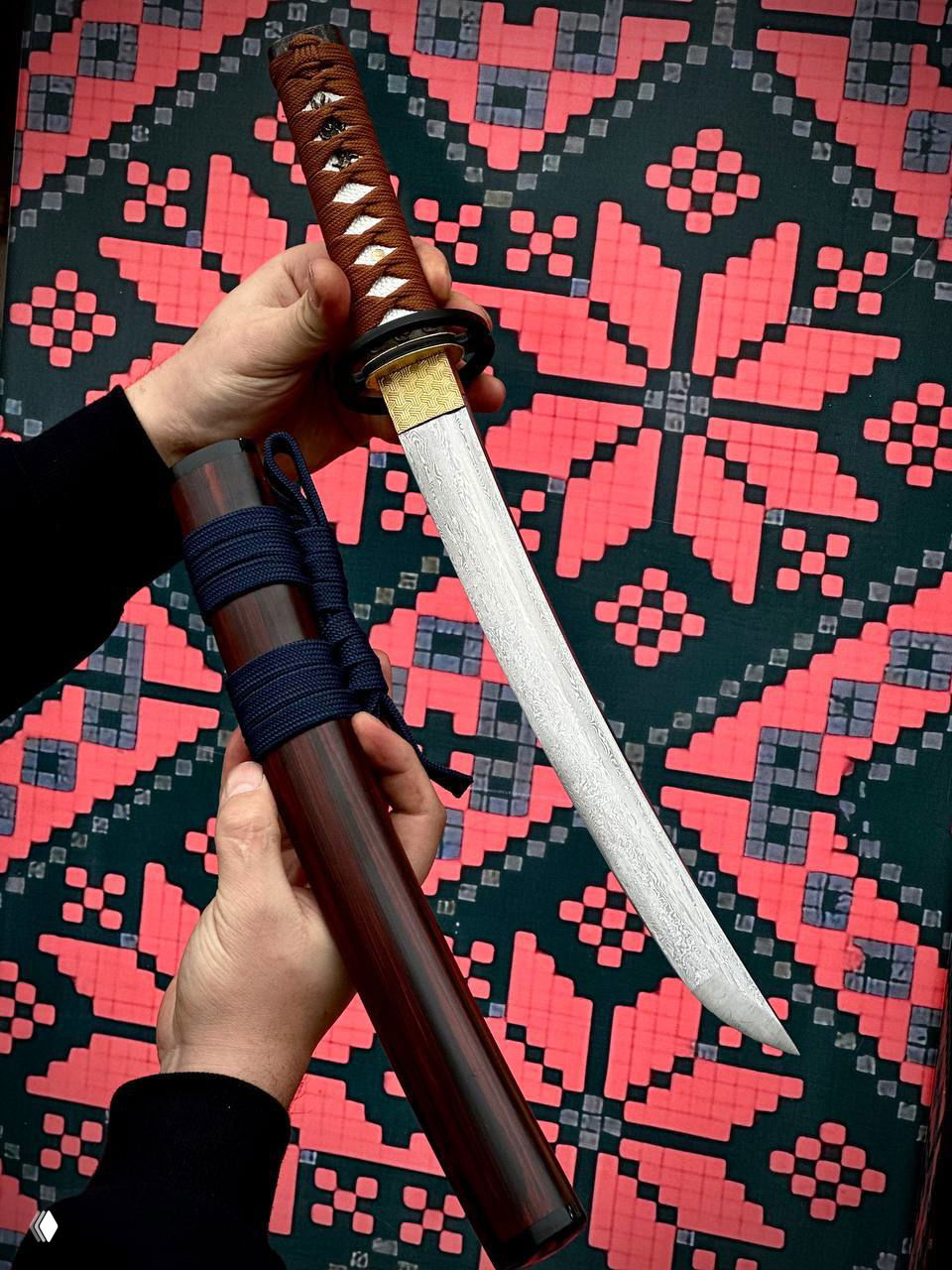 Руки демонстрируют вакидзаси с фигурным дамасским рисунком на лезвии и тёмно-коричневыми ножнами и обмоткой на рукояти.