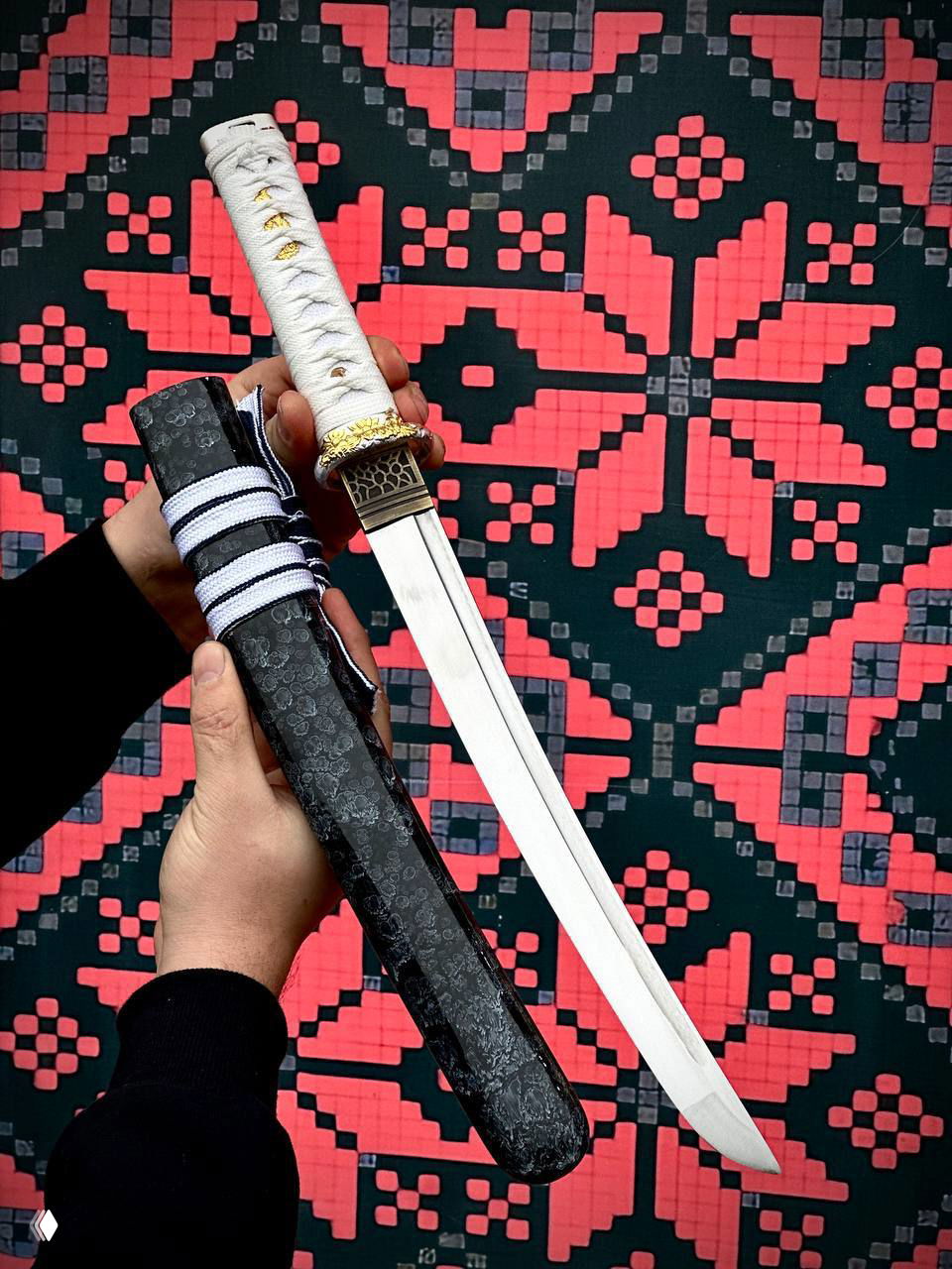 Белая оплётка рукояти вакидзаси и тёмные ножны в руках автора; хорошо видна граница клинка и цуба на фоне орнамента.