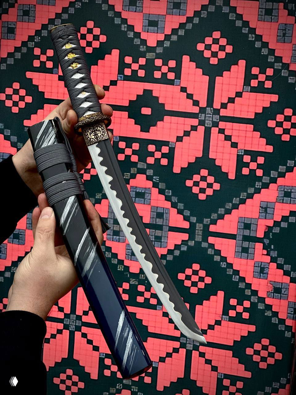 Длинный изогнутый клинок вакидзаси с тёмными ножнами в руках мастера, контрастный узор на фоне подчёркивает форму лезвия.