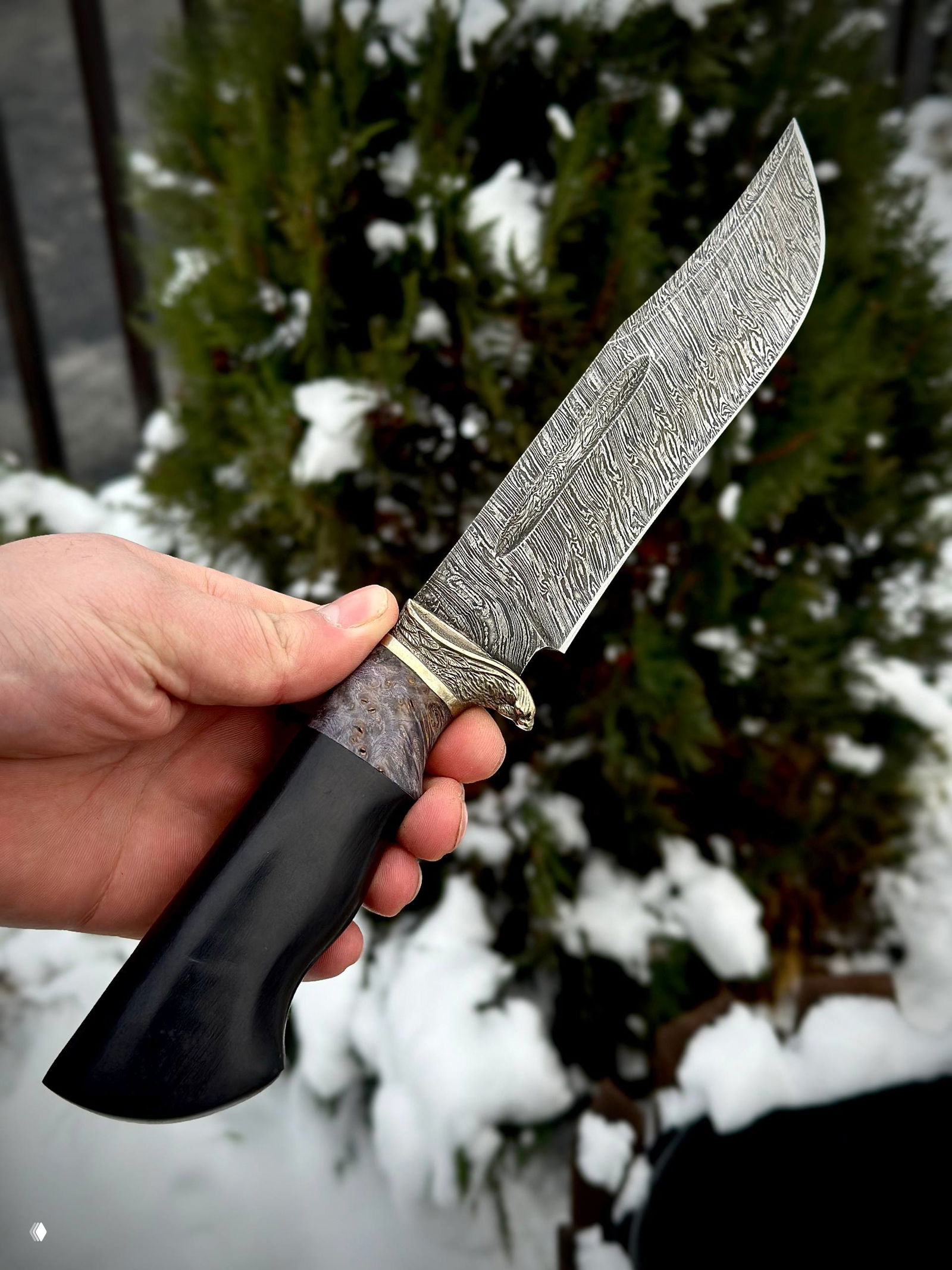 Фото клинка под углом: видно профиль лезвия и рисунок дамаска, а также деталь гарды и рукояти на фоне заснеженных ветвей.