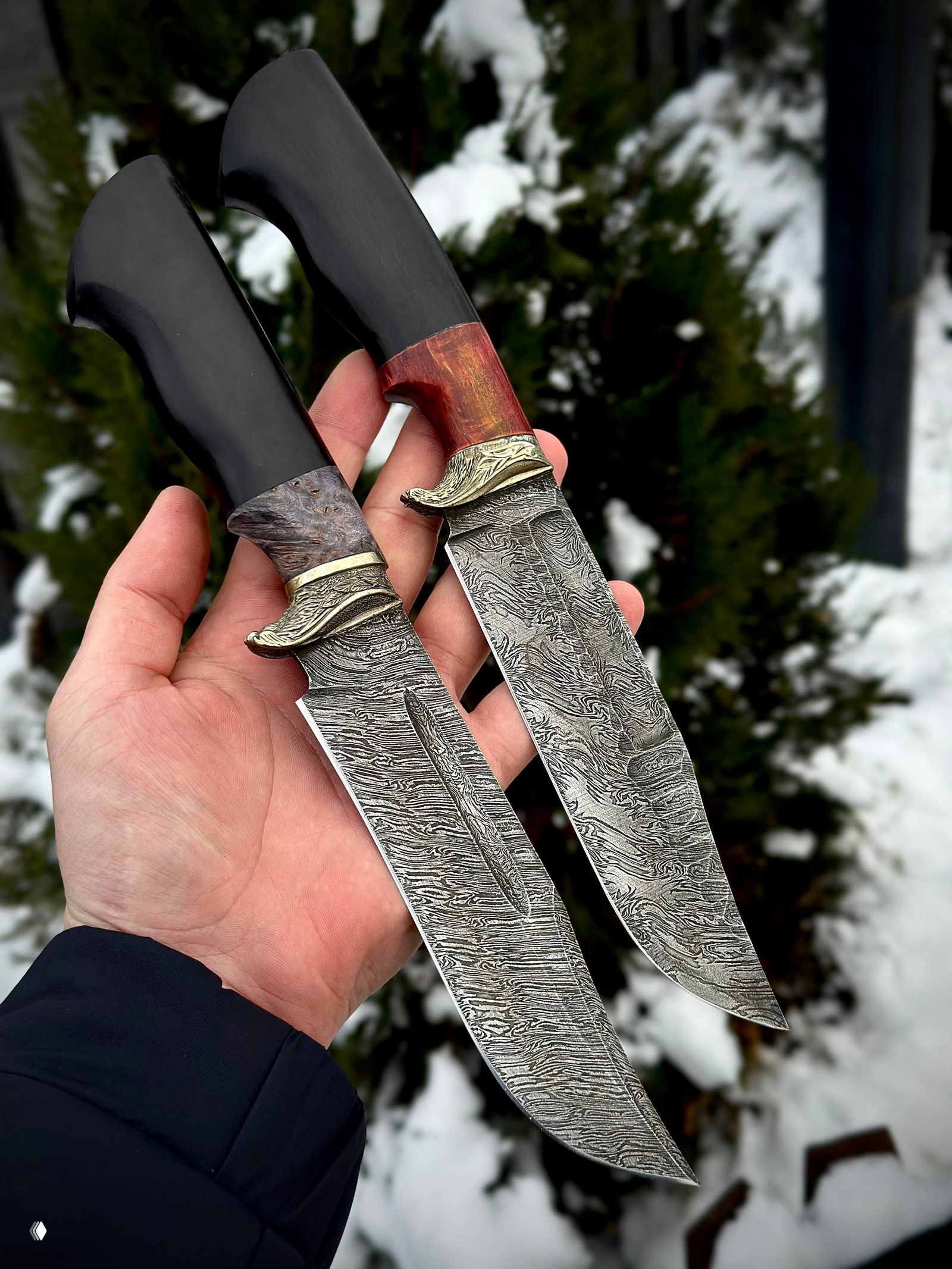 Два ножа в руке рядом: видно сравнение форм клинков и рукоятей, дамасская текстура на обоих лезвиях и украшенные гарды.
