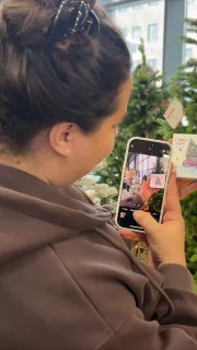 Покупательница снимает ёлку на смартфон в зоне продаж; на заднем плане видны другие модели и ценники на хвойных изделиях.