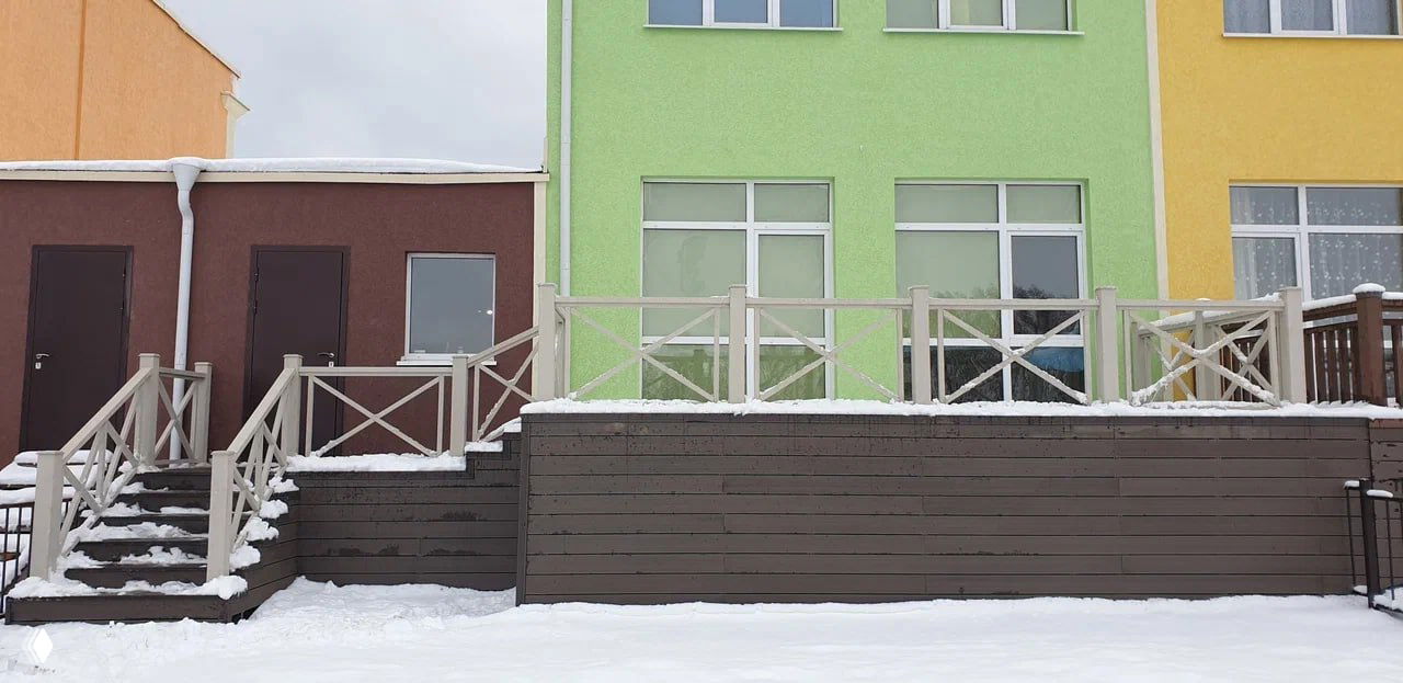 Терраса из ДПК с лестницей и перилами, снег на ступенях и настиле; на заднем плане фасад здания зелёного и жёлтого цветов.