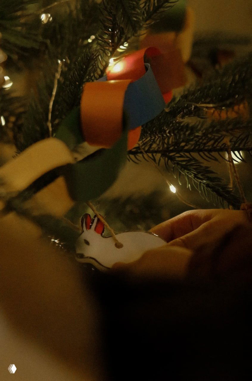 Рука взрослого или ребёнка вешает небольшую белую игрушку-зайчика на ветку ёлки рядом с цветной бумажной гирляндой и огоньками.