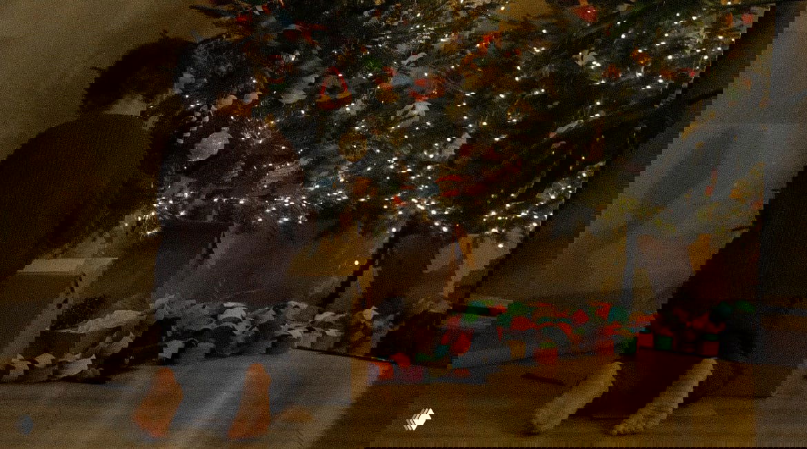 Широкий план нижней части украшенной ёлки, разноцветная бумажная гирлянда и коробки с подарками на полу в уютной комнате.