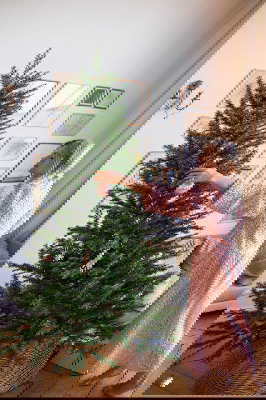 Полный вид: пышная искусственная ёлка «Уральская» 210 см в интерьере, женщина в розовом платье украшает ветви — фотография товара в жилой обстановке.