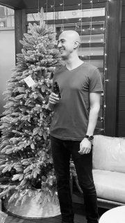 Чёрно-белое фото: мужчина (ведущий) стоит рядом с пушистой ёлкой в магазине, демонстрируя размер и внешний вид товара.