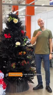 Чёрная искусственная ёлка с декоративными цветами и украшениями, рядом стоит ведущий в зелёной футболке в магазине.