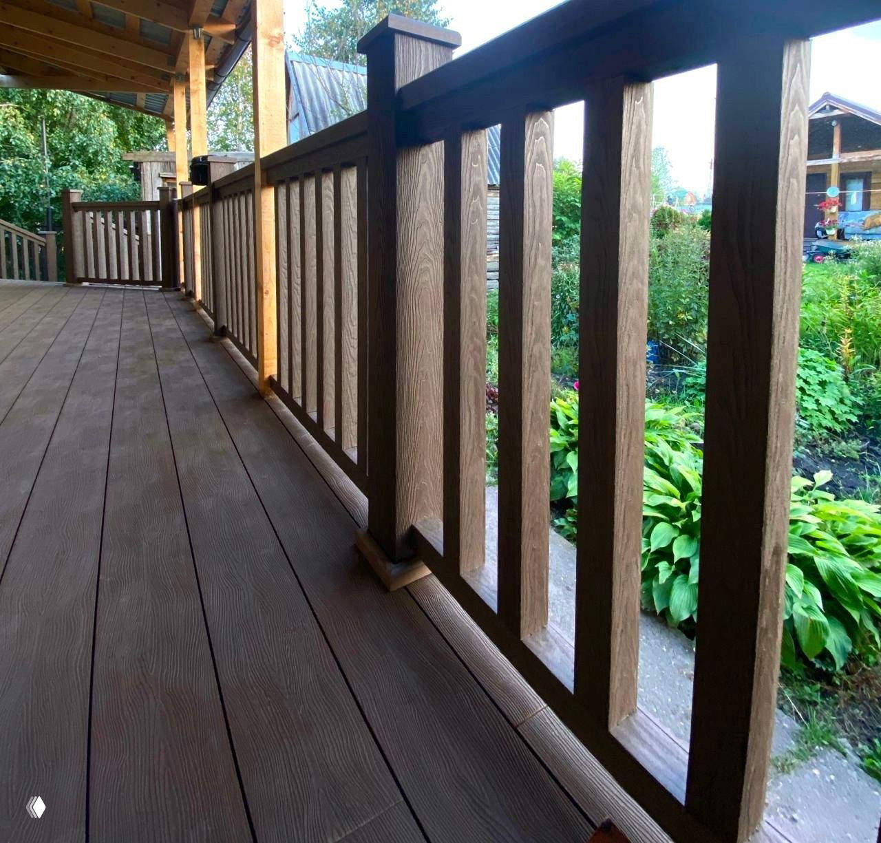 Фрагмент террасы с коричневой ДПК‑доской и ограждением, вид вдоль настила в перспективе, на фоне зелёного сада и дома.